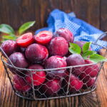 1-2 Tate IFF - Plum Flavour S - 3674 displayed as Fresh red plums in a rustic wire basket with green leaves and a blue cloth on wooden background