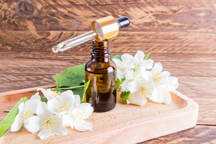 an open glass bottle of organic jasmine oil stands on a wooden tray with oil filled with a pipette. wooden brown background.