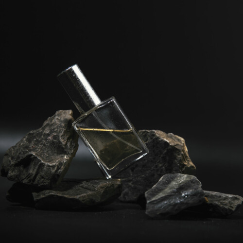1-2 Taste IFF - Rock Star Fragrance displayed in a sleek transparent glass bottle with a silver cap, placed on rugged black rocks against a dark background