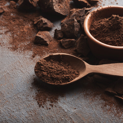 1-2 Taste JS Cocoa - Alkalized Cocoa Powder NERO™ UTZ MB shown as deep brown cocoa powder in a wooden spoon and bowl, surrounded by cocoa chunks on a rustic surface
