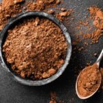 1-2 Taste JS Cocoa - Cocoa Powder ZERO™ displayed as a rich brown cocoa powder in a black bowl with a metal spoon, set against a dark textured background with scattered cocoa dust