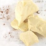 1-2 Taste JS Cocoa - Deodorized Cocoa Butter CURVE20™ UTZ MB displayed as creamy yellow cocoa butter chunks on a white wooden surface with cocoa powder flecks
