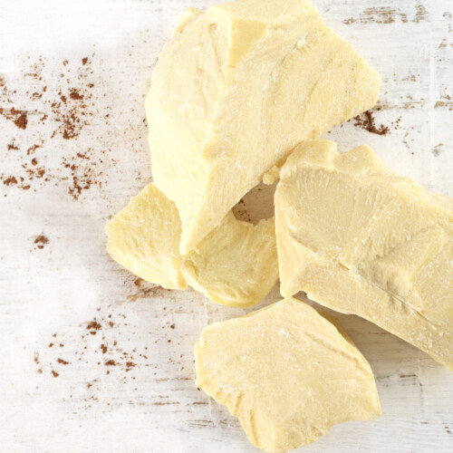 1-2 Taste JS Cocoa - Deodorized Cocoa Butter CURVE20™ UTZ MB displayed as creamy yellow cocoa butter chunks on a white wooden surface with cocoa powder flecks