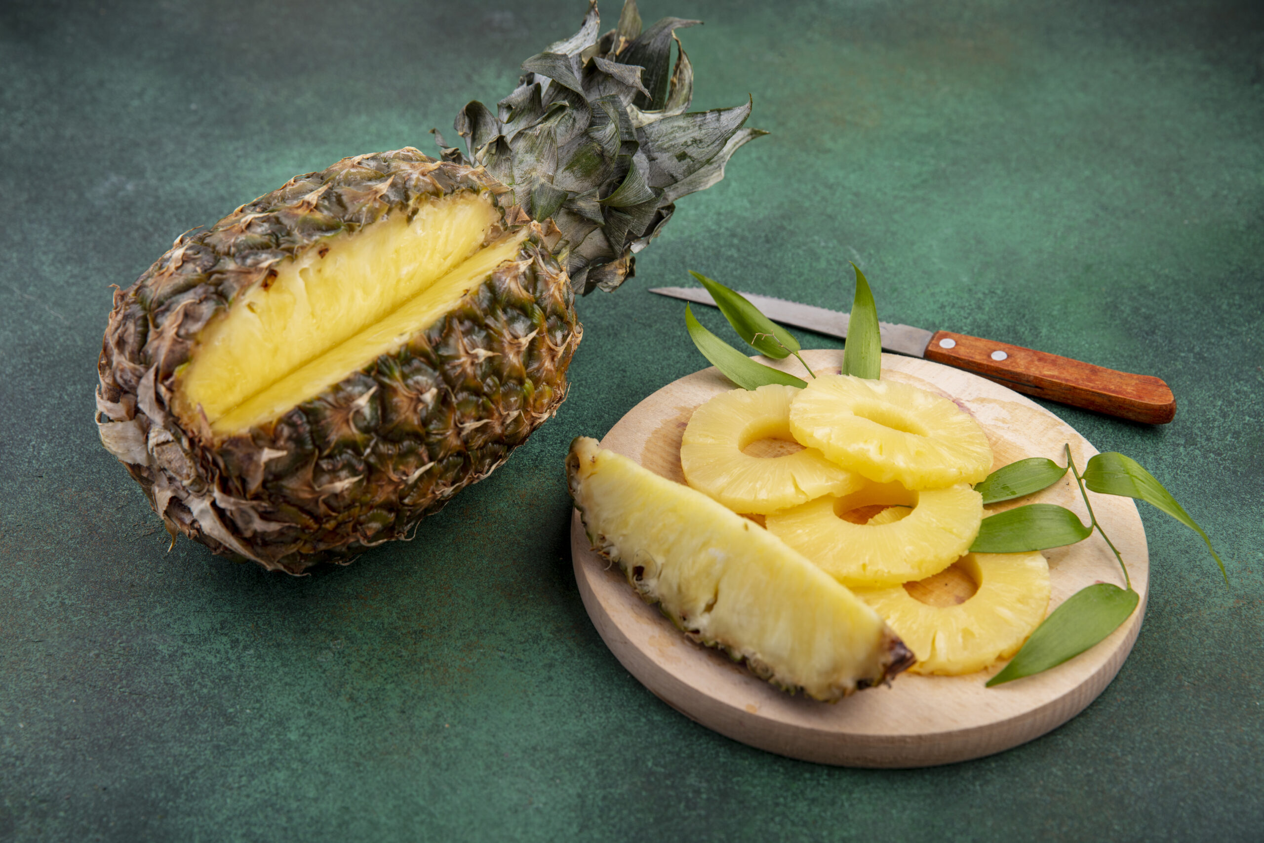side view of pineapple with one piece cut out from whole fruit and pineapple slices on cutting board with knife on green background