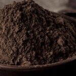1-2 Taste JS Cocoa – Mildly Alkalized Cocoa Powder UMBRA™, displayed as a heaped mound of rich brown cocoa powder in a dark ceramic bowl with soft lighting