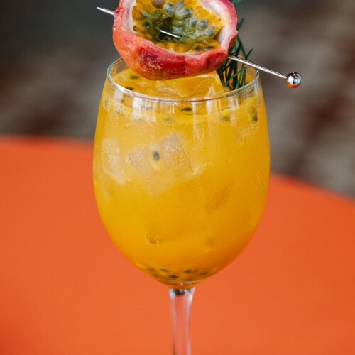 1-2 Taste Aroma INC - Natural Passionfruit Vanilla Flavour - O20231 shown as Tropical yellow drink with ice and passionfruit garnish in a wine glass on an orange surface