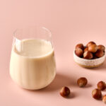 1-2-Taste Vegan Stock – Roasted Hazelnut Milk Base (RHPEU107.A9) shown as a glass of creamy hazelnut milk with whole roasted hazelnuts on a pastel pink background