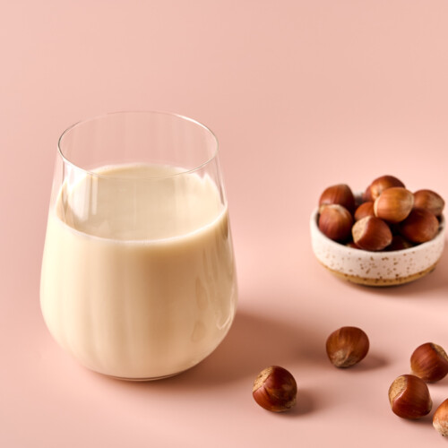 1-2-Taste Vegan Stock – Roasted Hazelnut Milk Base (RHPEU107.A9) shown as a glass of creamy hazelnut milk with whole roasted hazelnuts on a pastel pink background